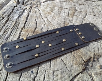 Könnte beinhalten: Dunkelbraunes Lederarmband mit messingfarbenen Nieten und Akzenten. Das Armband hat mehrere dünne Lederriemen und ein rechteckiges Design. Die Manschette wird mit Druckknöpfen und Nieten befestigt.