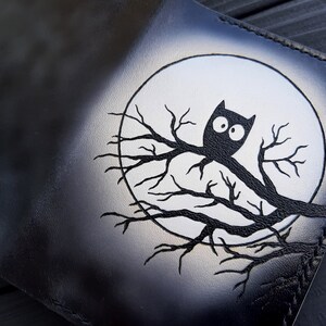 May include: Black leather wallet with a white moon and a black owl perched on a branch. The owl has large, white eyes.