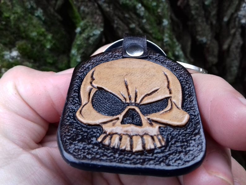 May include: A black leather keychain with a tan skull design. The skull is embossed on the leather, with detailed features including eye sockets, teeth, and a prominent brow. The keychain has a metal ring and a leather loop.