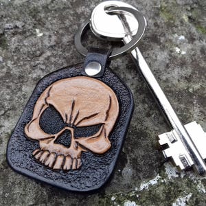 May include: A black leather keychain with a skull design. The skull is tan and embossed on the black leather. A silver key and key ring are attached to the keychain. The keychain is resting on a gray, textured surface.