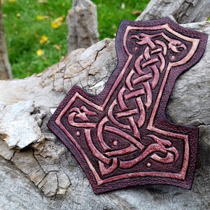 Puede incluir: Un parche de cuero con un diseño detallado de un Mjolnir, el martillo de Thor, en nudos celtas, de color burdeos con detalles dorados.