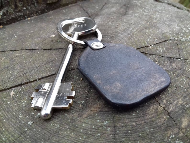 May include: A silver key and a black leather keychain rest on a weathered wooden surface. The key has a detailed design, and the keychain is a simple, rectangular shape, attached to a metal ring.