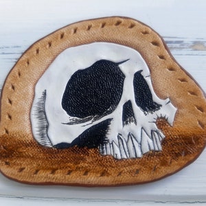 May include: A white leather skull patch with black eye sockets and teeth, stitched onto a brown leather background.