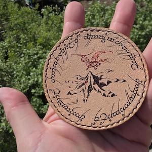Può includere: Un sottobicchiere rotondo in pelle marrone chiaro con una scena di montagna, un drago rosso e una scritta. Il sottobicchiere ha un bordo cucito ed è tenuto in mano.