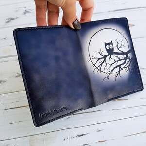 May include: A dark blue leather wallet with a white owl design on the front. The owl is perched on a branch in front of a full moon. The wallet has the text "Leather, Handmade" embossed on the bottom.