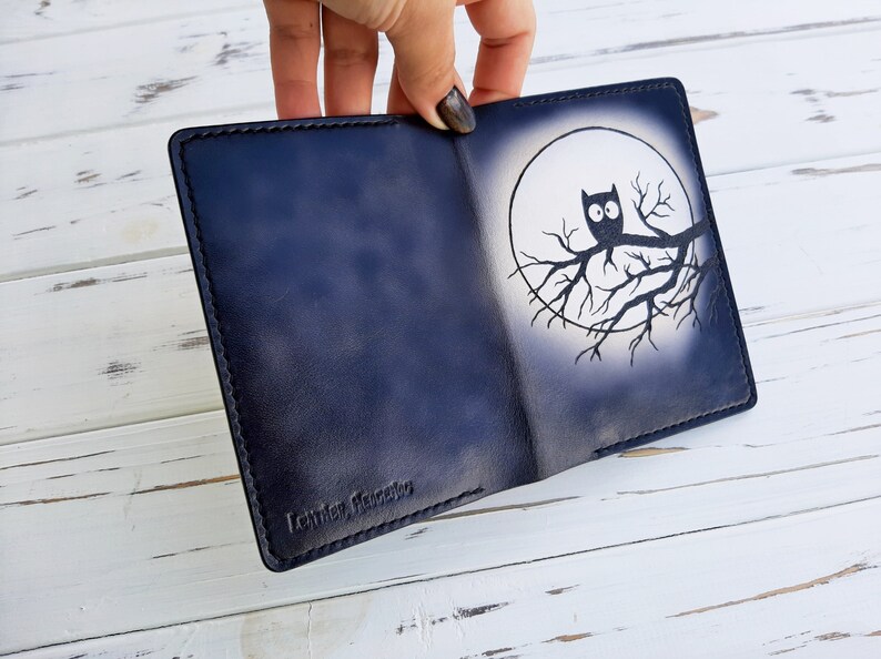 May include: A dark blue leather wallet with a white owl design on the front. The owl is perched on a branch in front of a full moon. The wallet has the text "Leather, Handmade" embossed on the bottom.