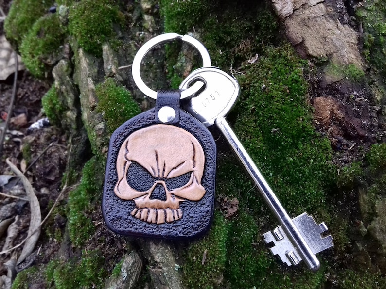 May include: A black leather keychain with a skull design in brown and tan. The keychain is attached to a silver key and a silver key ring. The background is a mossy tree trunk.