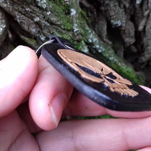 May include: Black and gold skull keychain. The keychain features a black, rectangular pendant with a gold skull design. The pendant is held by a black leather loop and a silver metal ring. The background is a blurred view of tree bark.