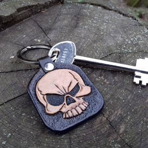 May include: A black leather keychain with a brown skull design. The skull is embossed on a rectangular tag, attached to a metal keyring. A silver key is next to the keychain, resting on a wooden surface.