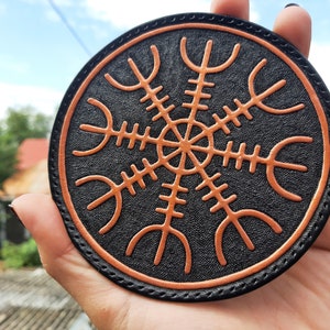 May include: A round leather coaster with a black background and an orange Vegvisir symbol. The coaster is stitched around the edge.