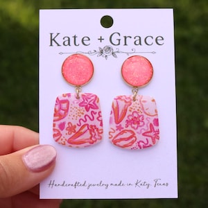 May include: Pink and gold earrings with a seashell and floral design. The earrings have a round top and a square bottom. The text "Kate + Grace" is at the top of the card. The text "Handcrafted jewelry made in Katy, Texas" is at the bottom.