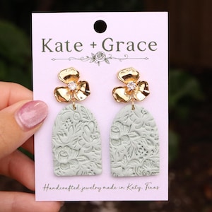 May include: A pair of handcrafted earrings with gold-colored flower tops and a clear gem center. The earrings have a light green, arched base with a floral pattern. The earrings are displayed on a pink card with the text "Kate + Grace".