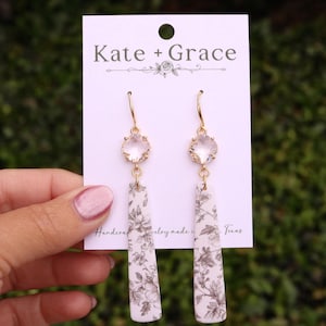 May include: Gold-tone earrings with clear, faceted crystal tops and rectangular, floral-patterned drops. The earrings are displayed on a white card with the text "Kate + Grace" and "Handcrafted jewelry made in Texas."