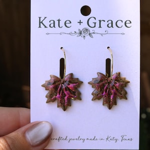 May include: A pair of hoop earrings featuring maple leaf-shaped charms with a glossy finish in shades of brown and metallic magenta. The earrings are displayed on a white card labeled Kate + Grace with the text handcrafted jewelry made in Katy, Texas.