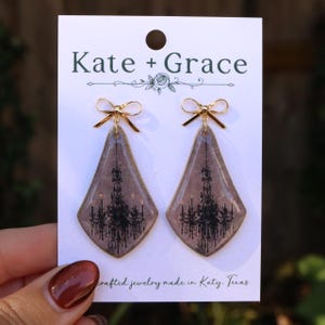 May include: A pair of diamond-shaped earrings on a white Kate + Grace display card. Each earring features a glossy, muted purple surface with a black chandelier graphic, topped with a small, polished gold-tone metal bow accent.