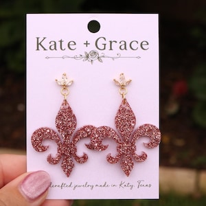 May include: Rose gold glitter fleur-de-lis earrings with a small crystal accent at the top. The earrings are hanging on a white card with the text "Kate + Grace" and "dercrafted jewelry made in Katy, Texas".