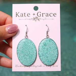 May include: Oval-shaped turquoise earrings with a glittery texture, hanging from silver-colored hooks. The earrings are displayed on a pink card with the text "Kate + Grace" and "Handcrafted jewelry made in Katy, Texas."