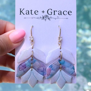 May include: Dangle earrings on a Kate + Grace display card. Each earring features a gold-tone hook with a clear crystal, followed by three layered leaf-shaped charms. The top leaf is white, the middle has a blue and purple pattern, and the bottom is white.