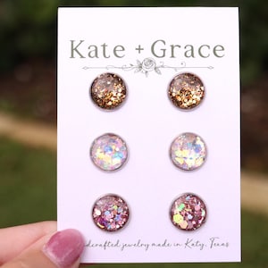 May include: Six pairs of round stud earrings with glitter and iridescent accents. The earrings are arranged on a white card with the text "Kate + Grace" and "dercrafted jewelry made in Katy, Texas". The earrings have silver-colored posts.