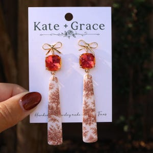 May include: Dangle earrings on a Kate + Grace display card. Each earring features a gold-tone metal bow at the top, a square orange faceted crystal, and a long, flat, rectangular pendant with a brown and white toile landscape print pattern.