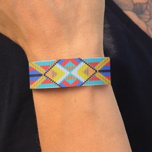 以下が含まれることがあります： 青、黄、赤、白の色合いで幾何学模様が織り込まれたカラフルな織りブレスレット。ブレスレットは手首に着用されています。