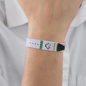 May include: A white woven bracelet with black, green and orange geometric patterns. The bracelet is on a person's wrist.