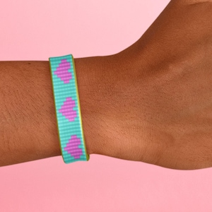 May include: A turquoise and green woven bracelet with a pattern of pink hearts. The bracelet is worn on a person's wrist.