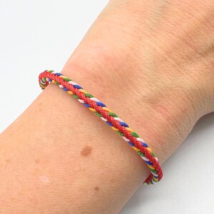 May include: Braided bracelet featuring red, white, blue, yellow, and green threads. The colorful bracelet is worn on a wrist, showcasing its vibrant design. This accessory adds a pop of color and style.