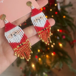 Può includere: Un paio di orecchini a forma di Babbo Natale in perle rosse e bianche con dettagli dorati. Gli orecchini sono pendenti e hanno una frangia di perle.