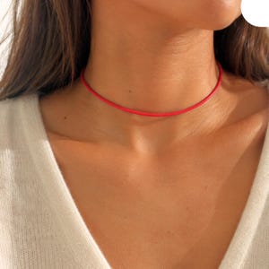 May include: A bright red choker necklace. The thin, round necklace sits close to the neck, creating a minimalist and modern look. The vivid red colour provides a pop of colour against the skin and a white sweater.