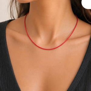 May include: A minimalist necklace in a vibrant red. The thin, round cord sits against the skin. The necklace is a solid red colour, offering a simple and elegant aesthetic.