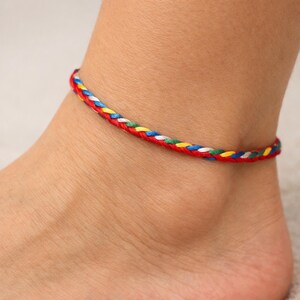 May include: Braided anklet featuring a vibrant mix of red, blue, white, yellow, and green threads. The colorful accessory is designed to be worn around the ankle. This anklet is a stylish piece of jewelry.