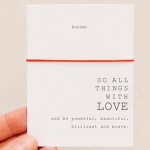 Può includere: Una carta bianca con il testo "DO ALL THINGS WITH LOVE" e le parole "and be powerful, beautiful, brilliant and brave." Un filo rosso è avvolto attorno alla carta. La parola "-bracelet-" è in alto.