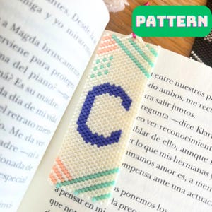 Op de afbeelding: Een witte kralenboekmarkeerder met een blauwe letter "C" in het midden. De boekmarkeerder heeft een patroon van roze, groene en witte strepen.