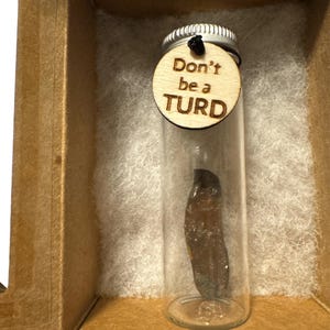 May include: A small glass vial containing a dark brown object, with a wooden tag that reads "Don't be a TURD." The vial is inside a cardboard box with white padding.