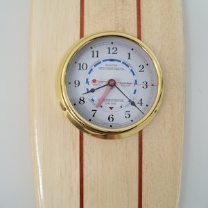 Puede incluir: Un reloj de madera con forma de tabla de surf con bisel de latón y esfera blanca. La esfera del reloj tiene números y manecillas negros, y un indicador de marea azul y rojo. El reloj está montado en una tabla de surf de madera con dos rayas rojas.