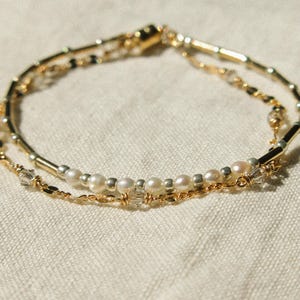 May include: A multi-strand bracelet featuring a combination of gold-toned metal tubes, small white pearls, and clear faceted crystal beads. The design incorporates delicate gold-colored chain links and wire-wrapped accents throughout the piece.