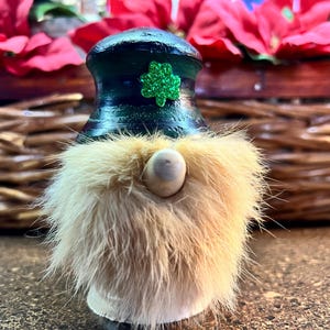 May include: A St. Patrick's Day gnome figurine with a green hat and a shamrock. The gnome has a tan fur beard and a wooden nose. The figurine is set against a blurred background of red flowers and a wicker basket.