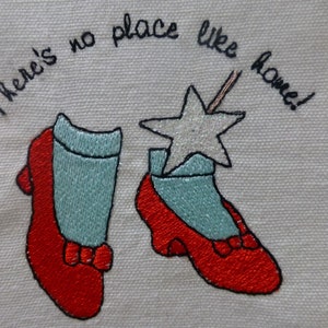 May include: Embroidered red shoes with a blue sock and a silver star on a white background. The text "There's no place like home!" is stitched above the shoes.
