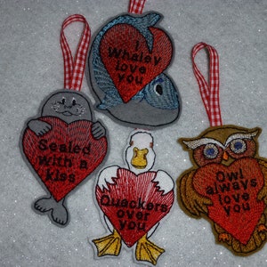 May include: Four felt ornaments with red hearts and animal designs. The ornaments feature a whale, a seal, a duck, and an owl. Each heart has a different message: "Whaley love you", "Sealed with a kiss", "Quackers over you", and "Owl always love you".