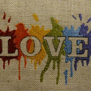 May include: Embroidered burlap fabric with the word "LOVE" in white block letters. The word is surrounded by colorful paint splatters in red, orange, yellow, green, blue, and purple. The background is a neutral beige.
