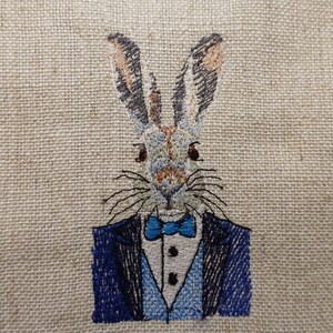 May include: Embroidered image of a rabbit wearing a blue suit and a blue bow tie on a beige linen background.