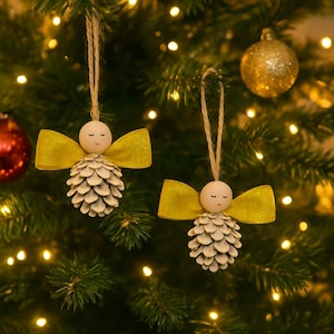 Puede incluir: Dos adornos navideños hechos a mano con forma de ángeles. Cada ángel tiene una cabeza de cuentas de madera con los ojos cerrados, un lazo de cinta amarilla y un cuerpo de piña. Cuelgan de un lazo de cuerda natural.