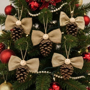 May include: Christmas tree ornaments featuring pinecones with burlap bows and wooden bead faces. The ornaments are suspended from twine and are decorated with red and gold Christmas balls. The tree is decorated with white pearl garland.