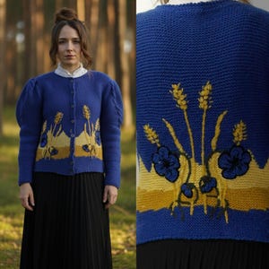 May include: A blue knitted cardigan featuring a floral and wheat design in yellow and black. The cardigan has a button-up front and puffed sleeves. The woman in the image is wearing a white collared shirt and a black skirt.