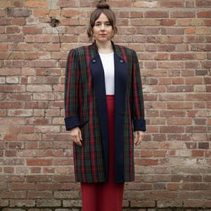 Può includere: Un cappotto lungo a quadri rosso, verde e blu navy, indossato sopra un top bianco e una gonna rossa. Il cappotto presenta un colletto e polsini blu navy, con due bottoni decorativi. L'outfit è davanti a un muro di mattoni.