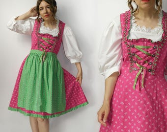 May include: A traditional pink dirndl dress with a green apron. The dress features a white blouse with puffy sleeves and a lace-trimmed neckline. The bodice is laced with green ribbon, and the skirt has a floral pattern. A classic Bavarian outfit.
