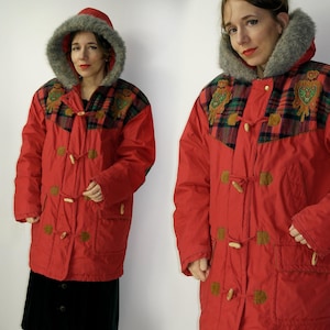 Op de afbeelding: Rode winterjas met een geruit en geborduurd ontwerp op de borst. De jas heeft een capuchon met imitatiebontvoering en knopen.