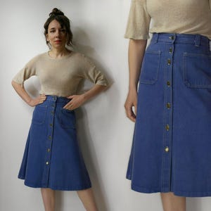 May include: A blue denim midi skirt featuring a high-waisted design with a full-length front button closure, two large patch pockets on the front, and visible contrast stitching. The skirt has a structured waistband and a flared silhouette.