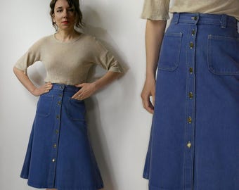 Op de afbeelding: Een blauwe denim midirok met een hoge taille, een volledige knoopsluiting aan de voorzijde, twee grote opgestikte zakken aan de voorkant en zichtbare contraststiksels. De rok heeft een gestructureerde tailleband en een uitlopend model.